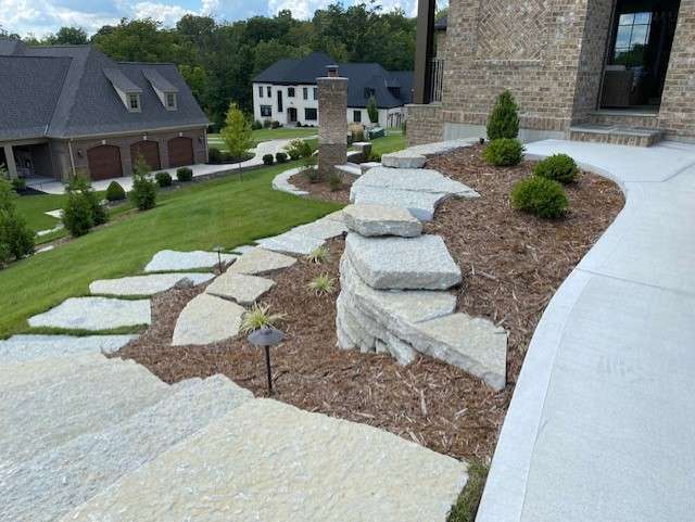 Stone slab steps ascend a landscaped mulch garden with small shrubs, highlighting the work of local landscaping companies beside a concrete walkway and brick house. Detached garages and neighbors' homes form the backdrop.