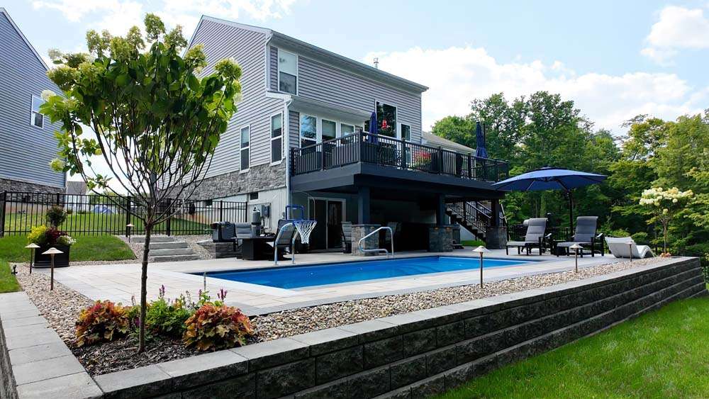 Modern two-story house with a raised deck overlooks a rectangular backyard pool, surrounded by patio seating, landscaping, trees, and elegant retaining walls for added structure and style.