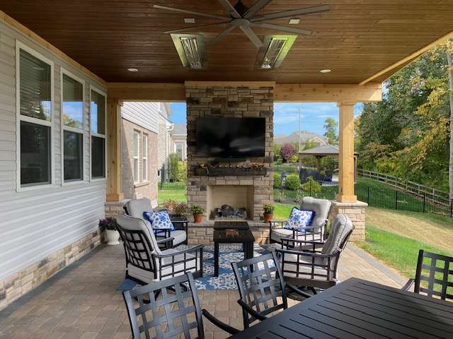 Covered patio with cushioned seating around a stone fireplace and TV, ceiling fans above, an outdoor dining table in the foreground, and backyard views—crafted by expert patio builders and landscaping companies for style and comfort.