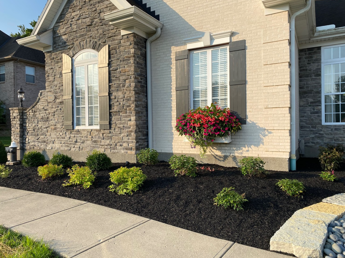 Front yard landscaping by expert masonry contractors features neatly trimmed shrubs, black mulch, and a window box of pink flowers on a brick house with stone accents.