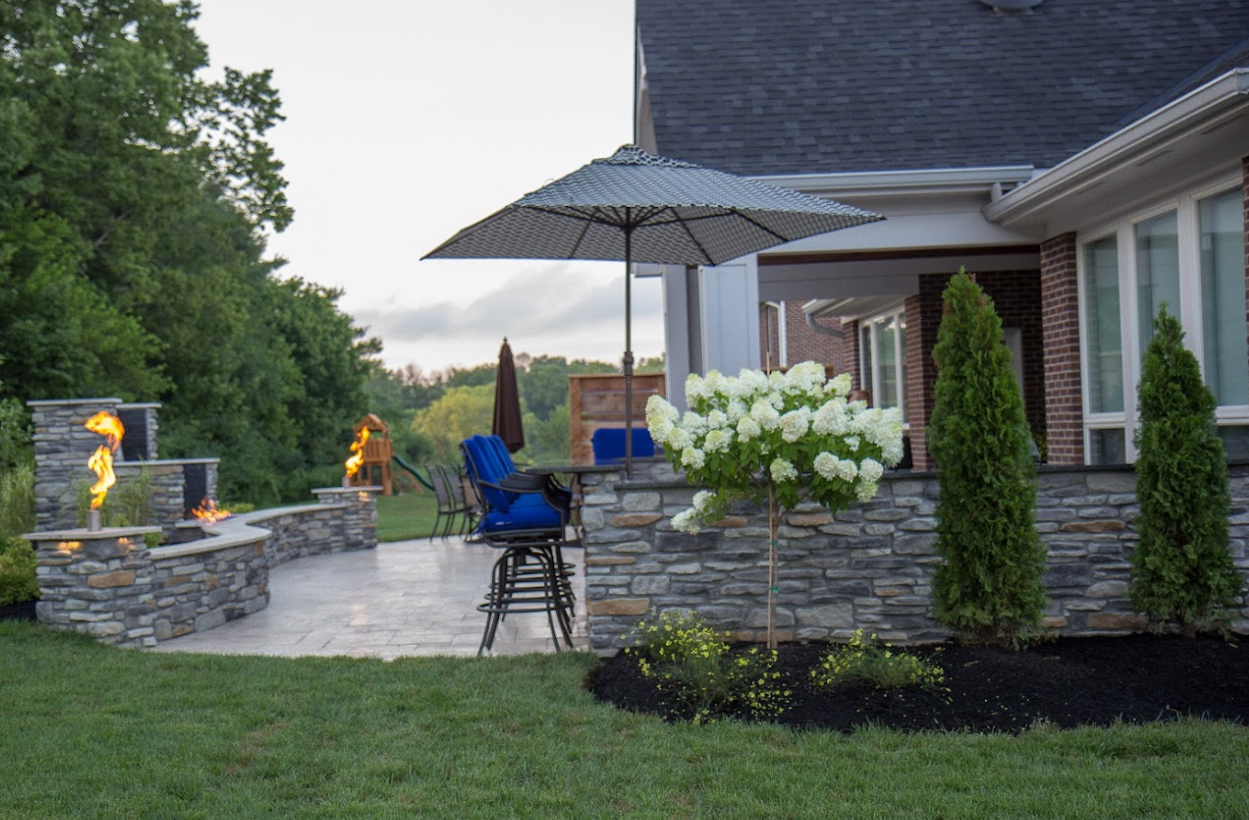 A backyard patio by leading patio builders features stone walls, fire pits, bar stools, a large umbrella, hydrangeas, and evergreens beside a brick house—perfectly landscaped and surrounded by lush lawn and trees.