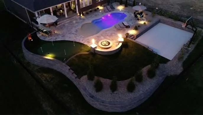 Aerial view of a landscaped backyard with a lit pool, fire pit, and patio at dusk.