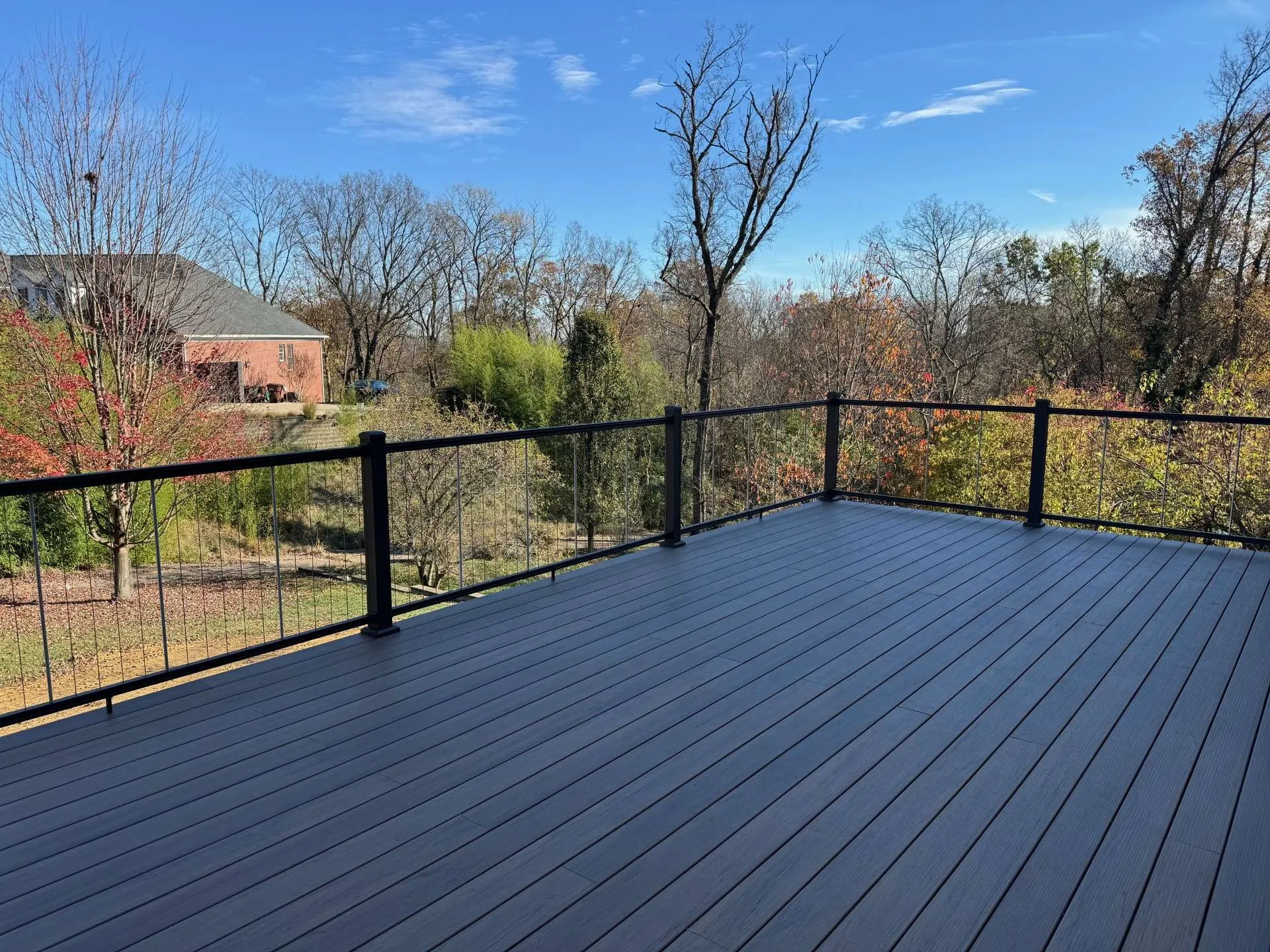 Gray composite deck with black cable railings overlooking trees, shrubs, and a neighboring brick house on a sunny day.