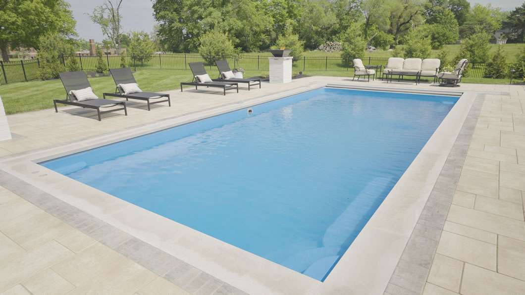 Rectangular outdoor swimming pool with clear water, surrounded by lounge chairs and patio furniture on a tiled deck, set in a grassy yard with trees in the background.