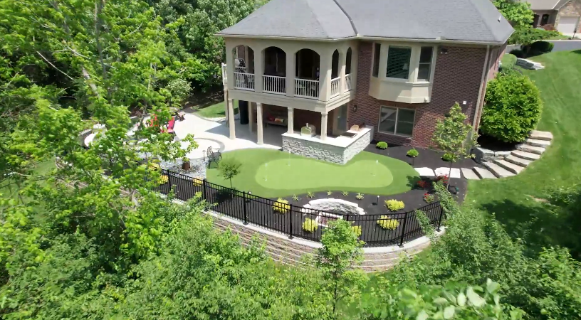 Aerial view of a two-story brick house with a covered patio, landscaped yard, and a small putting green surrounded by a black metal fence and lush greenery.